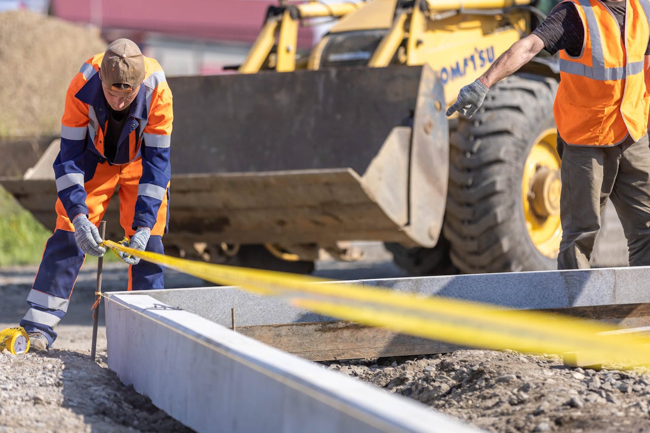 gallery-6 Construction workers using measuring tape at a sunny outdoor site with a bulldozer in the background.